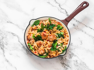 One pot lunch. Orzo pasta with meatballs and spinach in tomato sauce in a frying pan on a light background, top view