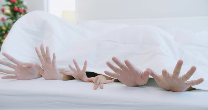 happy Asian family parent father mother and daughter lying on the bed hiding under white blanket and showing finger hand swing dance together with fun at bedroom home in the morning