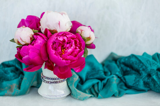 Bouquet of vibrant pink peonies and white peonies (Paeonia) in a white vase with decorative beads and a drape of teal colored fabric on a white counter top; Studio Shot
