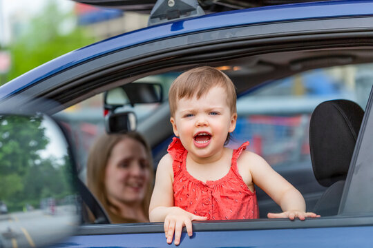 Toddler girl standing and looking out the window of a car with her mother sitting in the background; Burnaby, British Columbia, Canada