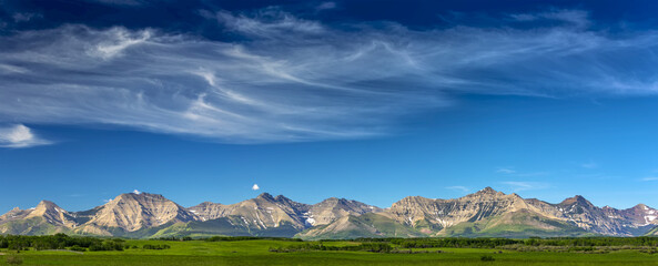 Panorama view of mountain range with wispy white clouds, blue sky and green field in the foreground, North of Waterton; Alberta, Canada