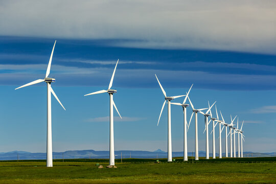 Rows of large wind turbines on a green field with clouds and blue sky and a mountain range in the distance, North of Waterton; Alberta, Canada