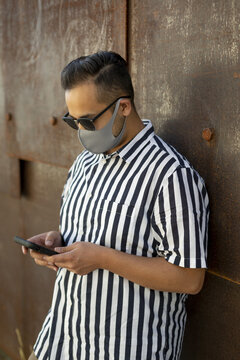 A man wearing a face mask and sunglasses stands looking down and using his smart phone, Lonsdale Quay; North Vancouver, British Columbia, Canada