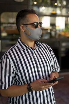 A man wearing a face mask and sunglasses stands looking out and using his smart phone, Lonsdale Quay; North Vancouver, British Columbia, Canada