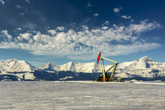 Pumpjack in a snow-covered field with dramatic clouds in a blue sky with snow-covered mountain range in the background; Longview, Alberta, Canada