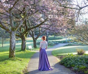 A woman with red curly hair and formal gown standing on a footpath in the gardens of a park; British Columbia, Canada