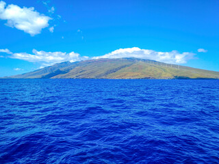Vibrant blue water of the Pacific Ocean and view of the West Maui Mountains with wind turbines lined up on its slopes under a bright blue sky; Maui, Hawaii, United States of America