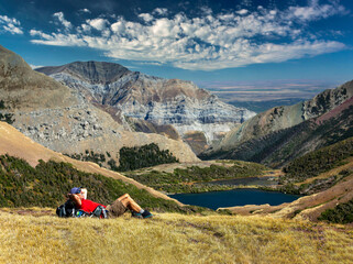 Male hiker relaxing on grassy mountain ridge overlooking an alpine lake, mountain ranges, blue sky and clouds in the background, Waterton Lakes National Park; Alberta, Canada