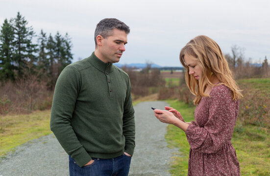 Mid-adult couple standing in a park, the wife using a smart phone as her husband watches her; Aldergrove, British Columbia, Canada