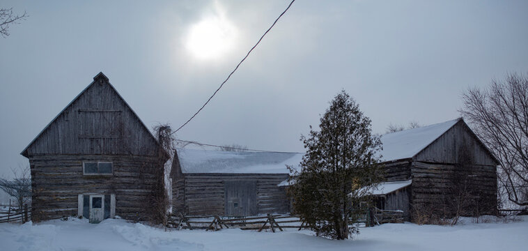 Weathered farm buildings in winter, with the sun glowing through the overcast sky; Ottawa Valley, Ontario, Canada