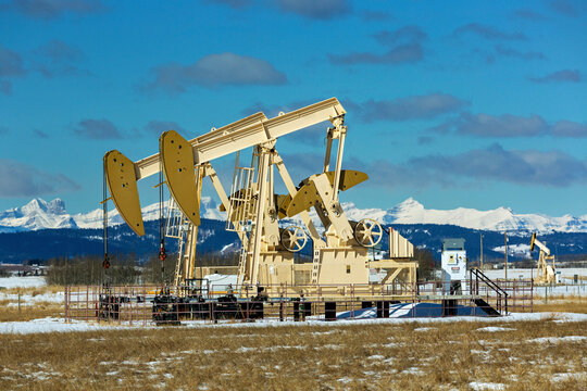 Pumpjacks  in a snow-covered field with snow-covered Rocky Mountains in the background with blue sky and clouds, West of Airdrie; Alberta, Canada