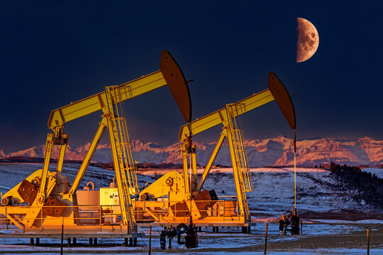 Colourfully warm lit pumpjacks at sunrise in a snow-covered field with glowing snow-covered mountain range and half moon in a deep blue sky, West of Airdrie; Alberta, Canada