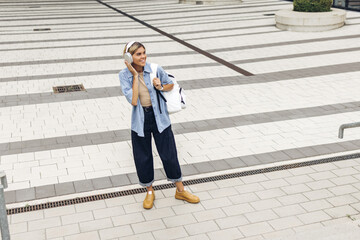 In full growth, positive young caucasian woman in good mood listens to music walking around city. Blonde wears shirt, jeans and backpack. Relaxed lifestyle, concept