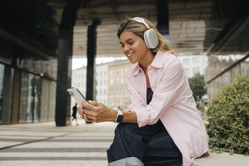 Positive caucasian young woman in headphones browsing playlist on phone sitting on street. Blonde...