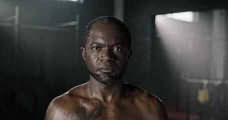Close-up portrait of serious confident strong African boxer looking at camera standing in gym. Shirtless muscular motivated African-American man professional mma fighter after training boxing.