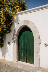 Fototapeta na wymiar Green wooden door with stone frame and white facade with yellow flowers, in Tavira, Algarve of Portugal.