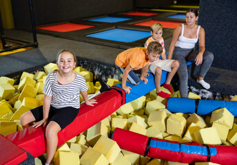 Group of happy tween children and young woman sitting on soft beam above pit of foam cubes,...