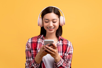 Happy asian lady with wireless headphones looking at smartphone and texting, posing on yellow background