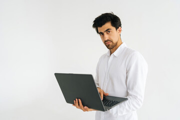 Studio portrait of serious confident young man holding in hand opened laptop computer and looking at camera standing on white isolated background. Pensive male posing with computer, copy space.