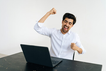 Studio shot of smiling excited male office worker screaming eureka raising hands up, solving hard task, getting access, successfully completing work sitting at desk on white isolated background. - obrazy, fototapety, plakaty