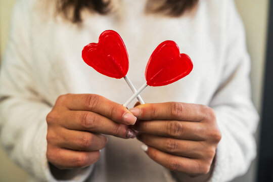 Close-up vertical image of a woman's hands dressed in white sweater crossing two red heart-shaped lollipops held in both hands. Young girl in love on Valentine's Day.