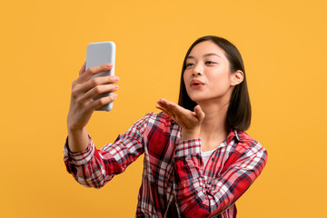 Joyful asian lady taking selfie with cellphone, sending air kiss and filming content for blog on yellow background