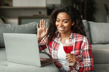 Glad millennial black lady waving hand, say hello greeting in computer camera, hold glass of wine...