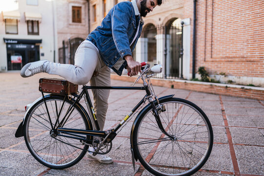 Cropped profile image of a young man in a denim jacket attempting to ride his vintage classic bicycle by swiping his leg over it.