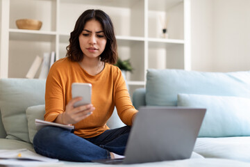 Young Middle Eastern Female Messaging On Smartphone And Using Laptop At Home