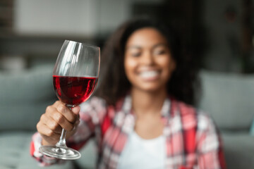 Smiling millennial black lady cheers glass of wine to camera in living room interior, blurred