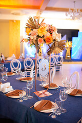 Festive table with blue cloth, napkins, plates, cutlery and glasses prepared for wedding guests in restaurant. Beautiful floral arrangement in gold metal vase with number three. Amazing servings