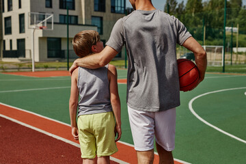 Back view of father and son playing basketball together and standing on court embracing