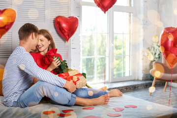 Young couple with red roses and gift for Valentine's Day in bedroom at home