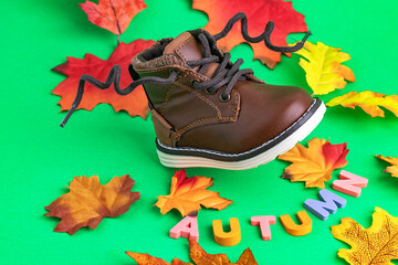 Leather, brown, children's boots with laces and autumn leaves on green background. The word AUTUMN from bright letters. Modern leather children's shoes.