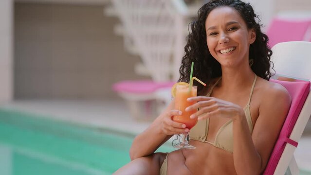 Sunbathing lady tanned female tourist traveler curly model arabian hispanic ethnic sexy woman drink cocktail fresh juice resting on poolside hotel spa flirting cheerful laughing enjoy summer vacation