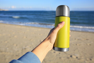 Woman holding yellow thermos with hot drink on beach near sea, closeup. Space for text