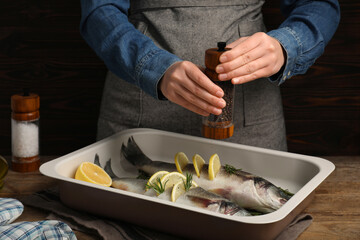 Woman grinding pepper onto raw sea bass fish with lemon and rosemary at wooden table, closeup