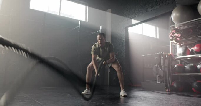 Attractive sporty fit young sportsman training in modern crossfit gym exercising with battle ropes. Handsome strong skinny male working out in shorts t-shirt and sneakers. Crossfit concept.