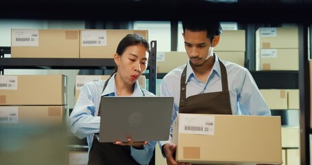 Asian business partner girl hold laptop and man wear formal shirt hold cardboard box training inventory online stock data program for delivery customer at warehouse. Startup small business concept.