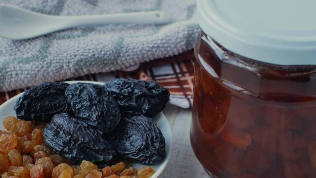 A jar of white cherry jam and a plate of dried fruits, raisins and prunes, a white glass teaspoon. Close-up side view from above. The concept of choosing healthy or unhealthy sugar