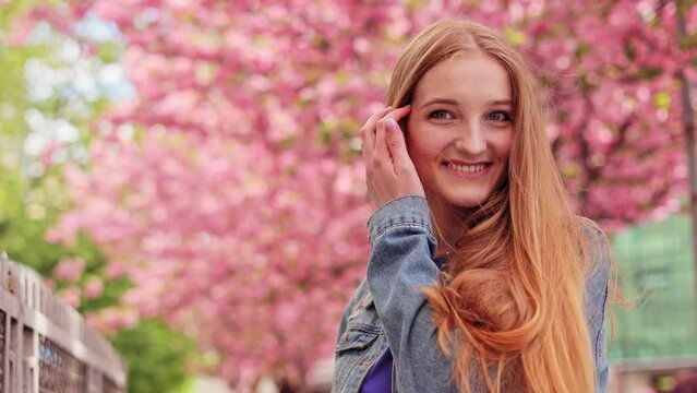 Happy Caucasian female with long hair in casual denim jacket walking around Cherry blossom trees. Portrait of young woman look around and smiling on blurred background in the city street. Sakura tree.