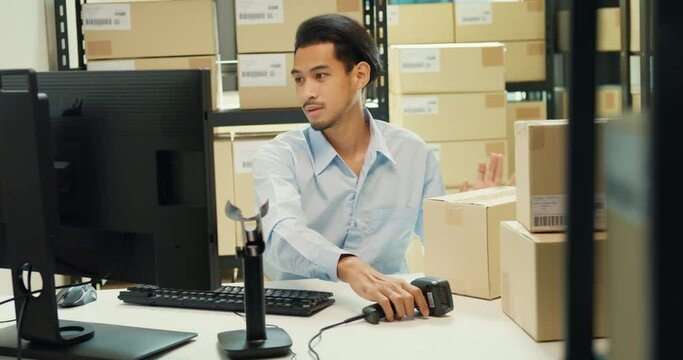 Happy Asian man wear formal uniform shirt sit in front of desk use computer and barcode machine scanner record customer online data in cardboard box delivery at warehouse. Startup business concept.