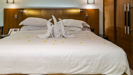 The bed is covered with white sheets. The blanket depicts a composition of folded towels in the form of a candle and a flame, yellow flower petals are scattered. The room is prepared for bed.