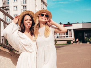 Naklejka premium Two young beautiful smiling hipster female in trendy summer white dress clothes and straw hats. Sexy carefree women walking in street. Positive models having fun, hugging and laughing