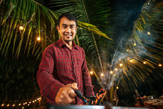 Portrait Of Happy Asian Young Man Cooking Meat On Barbecue Grill At New Year Party. Bar-B-Q Or BBQ On Traditional Stove. Night Party, People And Celebration Concept.