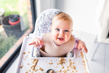 Happy six month old baby enjoying baby led weaning making mess on high chair tray