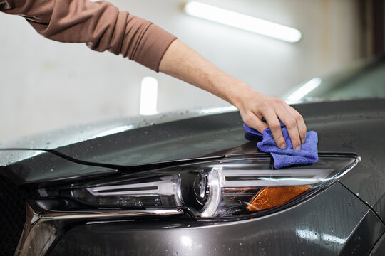 Closeup cropped image of hand of young man worker of auto service, wiping and polishing the headlights and hood of modern grey car with blue microfiber cloth. Car wash concept