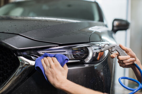 Closeup cropped image of hand of young man worker of auto service, wiping and polishing the headlights and hood of modern grey car with blue microfiber cloth under high pressure water.