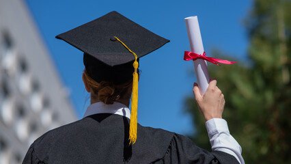 Rear view of a caucasian woman in a graduate gown holding a bundle with a diploma and standing near...