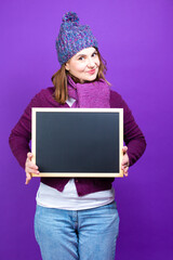 Modern Lifestyle Concepts. Positive Caucasian Female In Warm Knitted Winter Hat and Violet Scarf With Black Wooden Blackboard On Trendy Purple